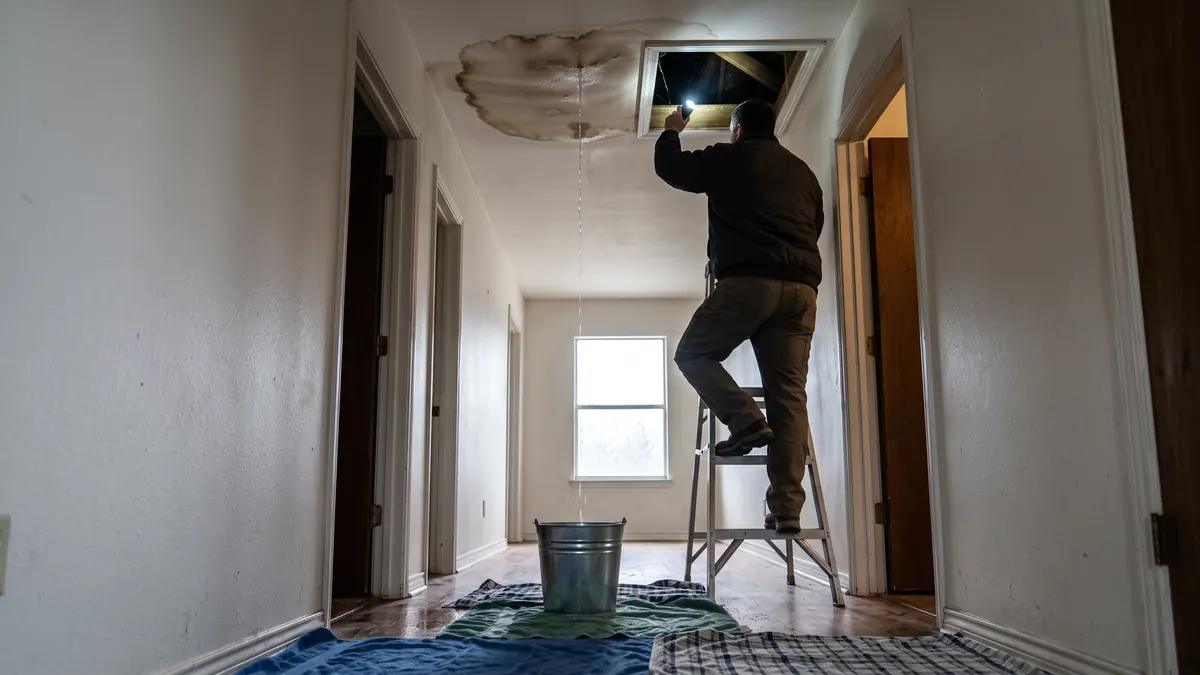 Technician on a step ladder inspecting a ceiling water stain below an attic access panel